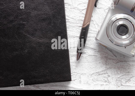 Top view of black notebook and pen near photo camera on white desk background for mockup. There is a place for your text. Stock Photo