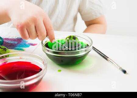 Child's hand holding spoon with green egg, child painting quail eggs for Easter on white table Stock Photo