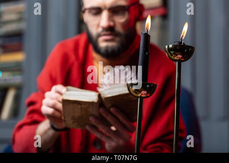Bearded diviner wearing glasses reading old book sitting near candles Stock Photo
