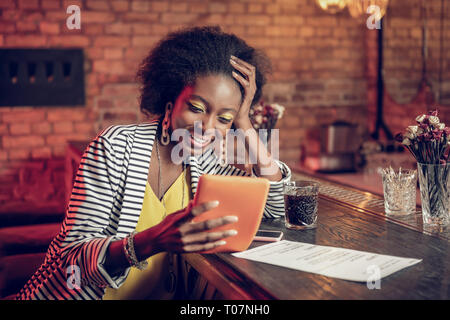 Bewitching Afro-American woman watching funny video on tablet at bar Stock Photo