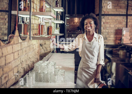 Smiling bewitching dark-haired African-American waitress standing near bar Stock Photo