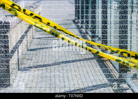 Broken and fractured Georgian wired glass panel with yellow caution safety tape across, installed along a public walkway with cement sidewalk in backg Stock Photo