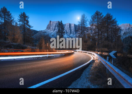 Blurred car headlights on winding road at night in autumn. landscape with asphalt road, light trails, mountains, trees, and blue sky with moon at dusk Stock Photo