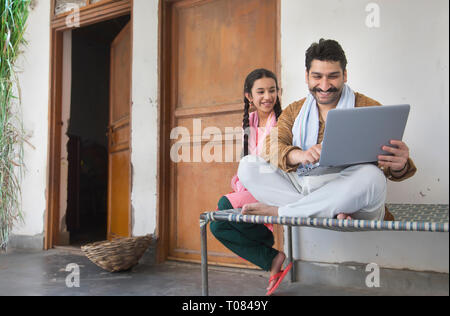 Happy village man sitting with her daughter on a cot operating a laptop computer. Stock Photo