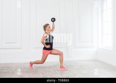 Side view portrait of young athletic beautiful bodybuilder woman in pink shorts and black top holding dumbells over head and doing squats with one leg Stock Photo