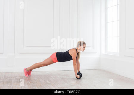 Full length side view portrait of athletic young woman in fit wear using dumbells in gym to workout and standing on perfect plank. Indoor, studio shot Stock Photo