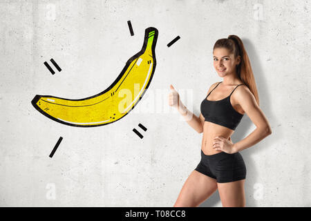 Young fit woman in black gym crop top and shorts standing in half-turn doing thumbs up pointing at drawing of big banana on wall. Stock Photo