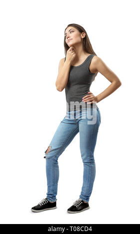 Young woman in sleeveless top and blue jeans standing in half-turn with left hand on hip and right arm rubbing under chin isolated on white background Stock Photo