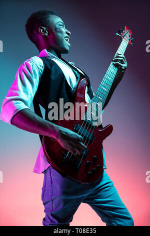 African American handsome jazz musician playing bass guitar in the studio on a neon background. Music concept. Young joyful attractive guy improvising. Close-up retro portrait. Stock Photo