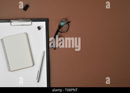 Flat lay, top view office table desk. Workspace with blank clip board, office supplies, eyeglasses, notepad and pen on a brown background. Stock Photo