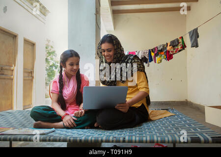 Happy village woman sitting with her daughter on a cot at home and operating a laptop computer. Stock Photo