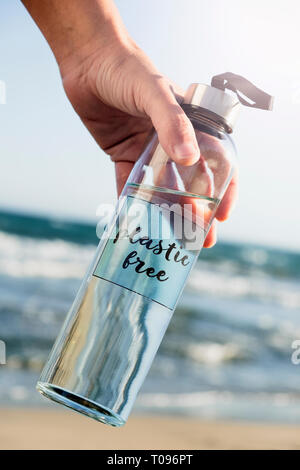 closeup of a caucasian man holding a glass reusable water bottle with the text plastic free written in it, on the beach, with the ocean in the backgro Stock Photo