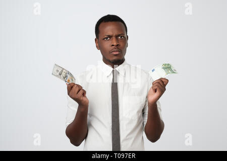 Young attractive african man holding USA currency and euro currency deciding which one to choose. Stock Photo