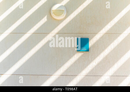 Outdoor light fixture and a blue no smoking sign on textured white wall and rays of sunlight in Umm Al Emarat Park, Abu Dhabi, United Arab Emirates Stock Photo