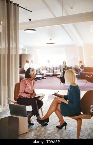 Two young work colleagues sitting together in an office discussing paperwork and using a laptop Stock Photo