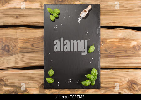 Slate server/board/tray with space for text on rustic wood planks. Arranged salt crystals, wooden scoop and basil leaves. Stock Photo
