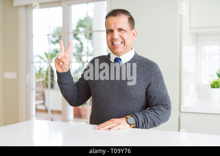 Middle age man sitting at home smiling with happy face winking at the camera doing victory sign. Number two. Stock Photo