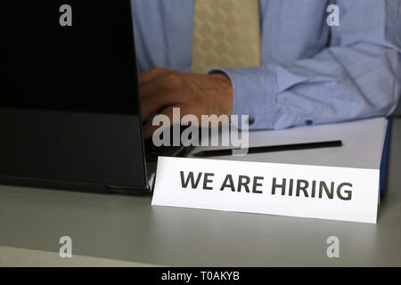 Hiring and recruitment, man in blue shirt and tie sitting at desk with laptop near the sign with the inscription We are hiring. Concept of headhunter Stock Photo