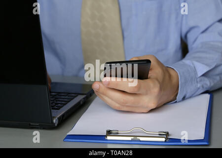 Office work, man in blue shirt and tie sitting at a table with laptop. Male hands typing on keyboard, using smartphone, concept of manager, clerk Stock Photo