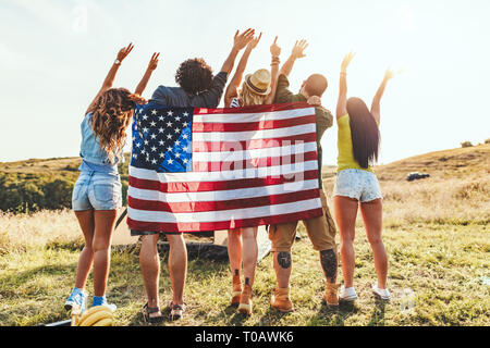 Happy young friends enjoy a sunny day in nature. They're looking at sun holding american flag and greeting, happy to be together. Stock Photo