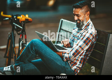 Smiling handsome young man is on a break. He is sitting on a bench and working at laptop, happy because of success, next the bench rests a bike. Stock Photo