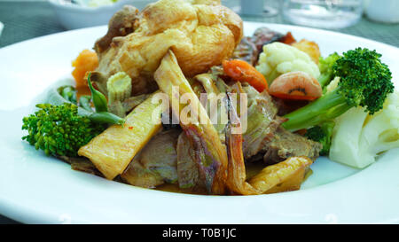 Traditional English Sunday lunch food of Yorkshire pudding, slices of meat, roast potatoes, carrots, broccoli, cauliflower, beans, served on a white p Stock Photo