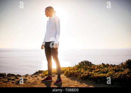 Young woman takes a break from her sunrise jog along the cliffs to admire the view of the ocean. Stock Photo