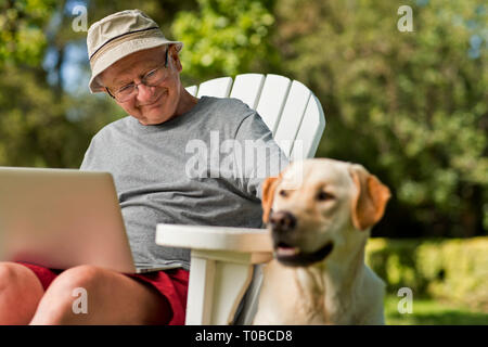 Senior man using his laptop in the garden, with his dog by his side. Stock Photo