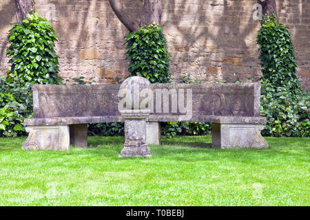 Grey stone bench by a wall with trees covered with ivy, in front of lawn, English garden on a summer day . Stock Photo