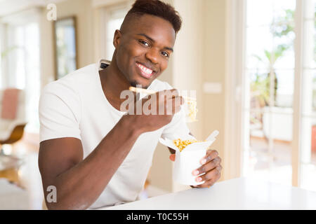 Handsome african man eating asian noodles in a delivery box, smiling enjoying lunch using chopsticks Stock Photo