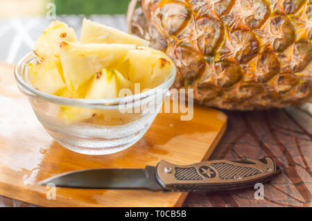 Close up side view of pineapple pieces and slices kept in glassware on a table besides a whole pineapple and a knife kept on chopping plate Stock Photo