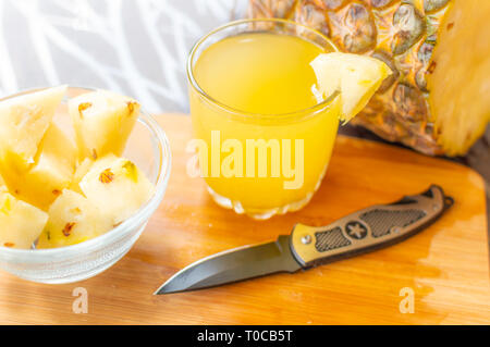 Side view of Sliced pineapple kept on a wooden table besides a glass filled with pineapple juice and a glass bowl with pineapple slices and a knife ke Stock Photo