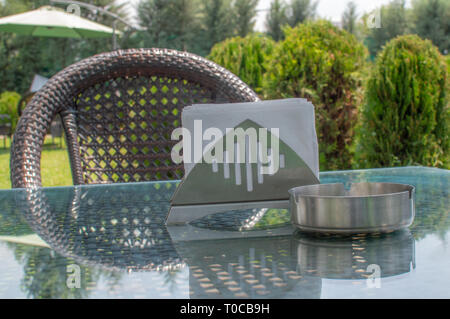 A chair with a table and ash tray on the glass table in a cafe with lush green plants Stock Photo