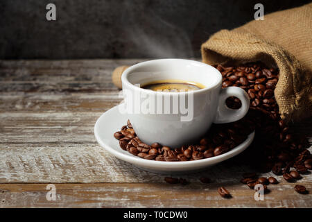 Coffee cup and roasted coffee beans in a burlap sack on a rustic wooden table on rusty vintage background. Stock Photo