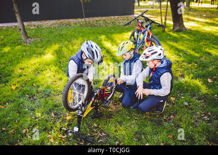 Teamwork. big family three Caucasian children brothers are learning to repair a bicycle, use tools in helmets same clothes, against of building up on Stock Photo