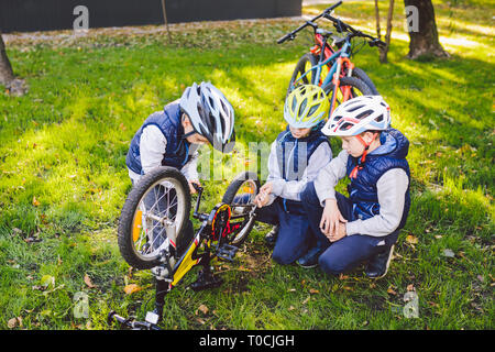 Teamwork. big family three Caucasian children brothers are learning to repair a bicycle, use tools in helmets same clothes, against of building up on Stock Photo