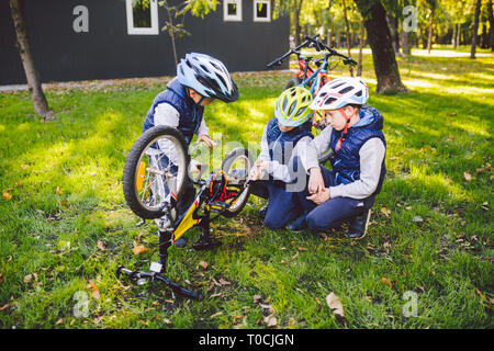 Teamwork. big family three Caucasian children brothers are learning to repair a bicycle, use tools in helmets same clothes, against of building up on Stock Photo