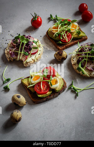 Healthy sandwiches with avocado, tomato, quail eggs, radishes and micro greens (sprouts) on a dark background, hard light Stock Photo