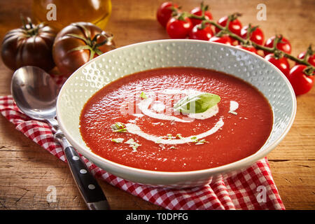 Plate of tomato cream soup with greens, served on red checked napkin on wooden table. Different kinds of tomatoes and jar of oil in background Stock Photo