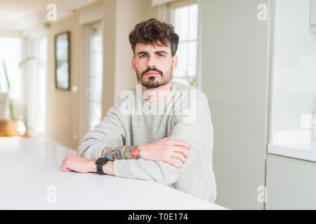 Young man wearing casual sweater sitting on white table skeptic and nervous, disapproving expression on face with crossed arms. Negative person. Stock Photo