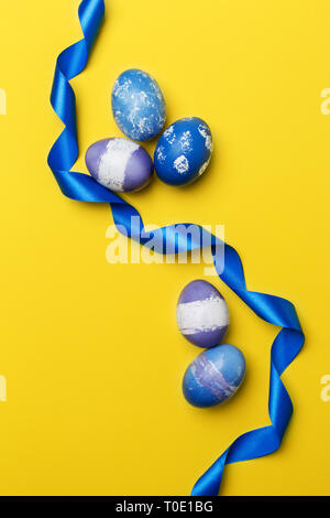 Festive Multicolored Easter eggs with blue ribbon on yellow background. Blue, purple and violet boiled chicken eggs with blue ribbon. Concept of Easte Stock Photo