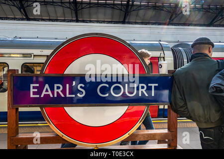 A man leans against station roundel sign on the platform at Earls Court Underground Station ,London, UK Stock Photo