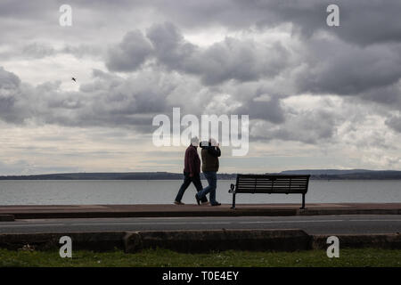 A middle aged couple walking along the promenade at the beach on a chilly and windy march day Stock Photo