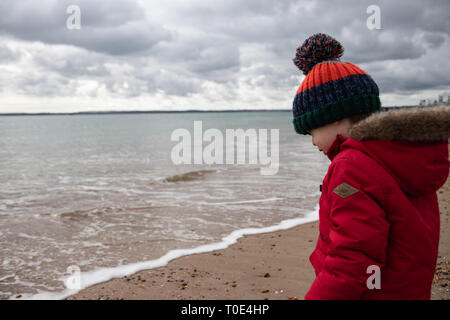 Young boy aged two in red coat playing on the beach in spring with bobble hat Stock Photo