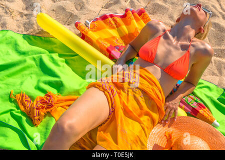 A woman tanning lying on the beach with clothes and equipment by sea bright orange, green and yellow colors. Among the objects: sarong, hat, swimsuit Stock Photo