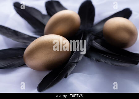 Three golden Easter eggs laying on a bunch of black feathers, on white linen as background Stock Photo