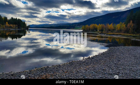 Peaceful and quiet untouched landscape scenery of pebble beach and lake with dramatic clouds reflected in the calm water with autumn trees around Stock Photo