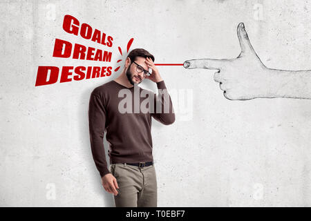 Young man standing half-turn, hand at forehead, at wall with drawing of big finger gun shooting words 'Goals, Dream, Desires' at him. Stock Photo