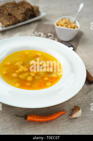 Chicken soup with vegetables in white bowl with croutons crackers and bread pepper on tablecloth. Stock Photo