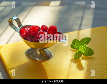 Ripe sweet raspberries in silver bowl and mint on yellow kitchen board in lights of sun. Close up, top view, healthy breakfast ingredient Stock Photo